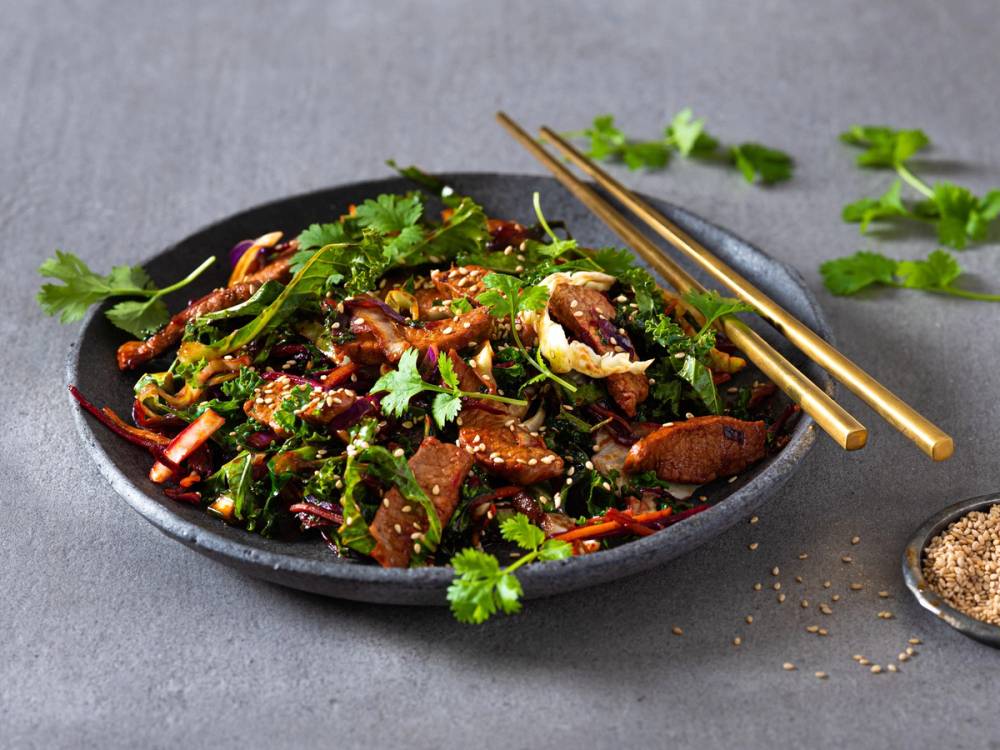 En wok med grønnsaker og kjøtt, pyntet med fersk koriander, på en svart tallerken med spisepinner ved siden av.