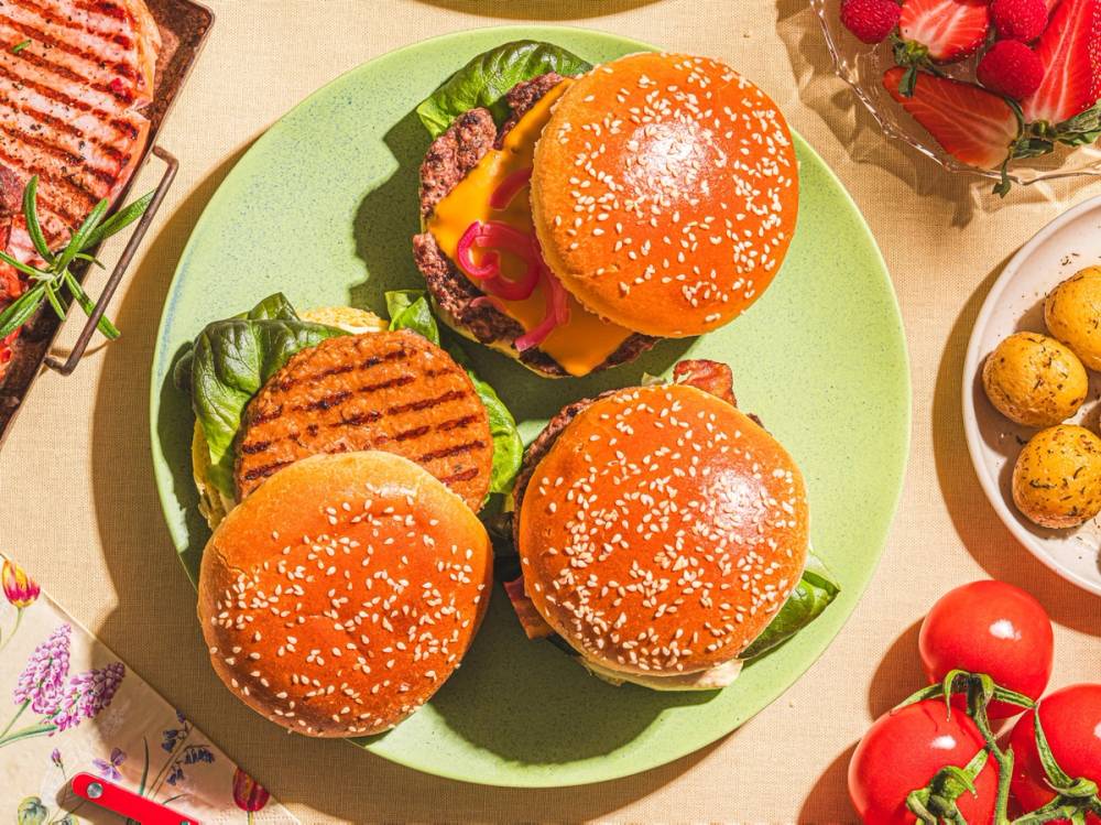 Tre hamburgere med brød, salat og kjøtt på en grønn tallerken, omgitt av små tomater og andre matvarer.