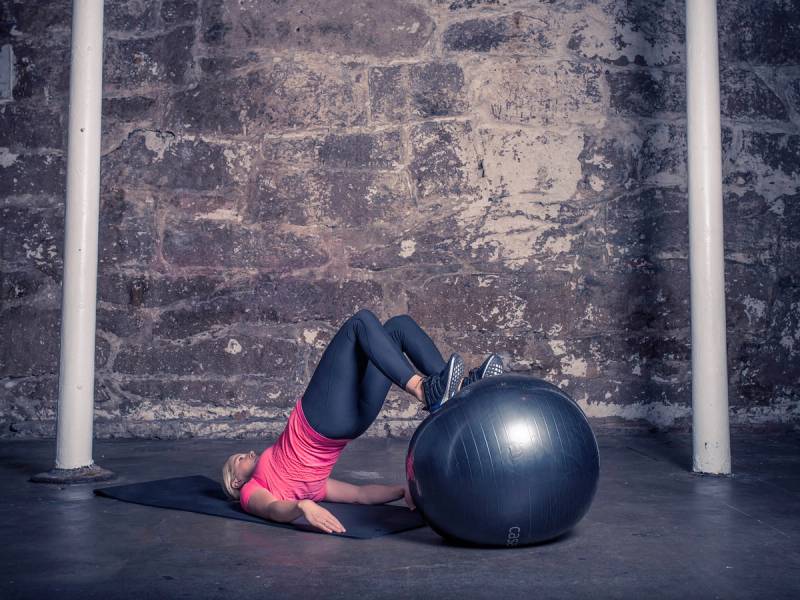 En person ligger på gulvet med beina oppå en stor treningsball i et treningsrom med murvegg og stangstativ.
