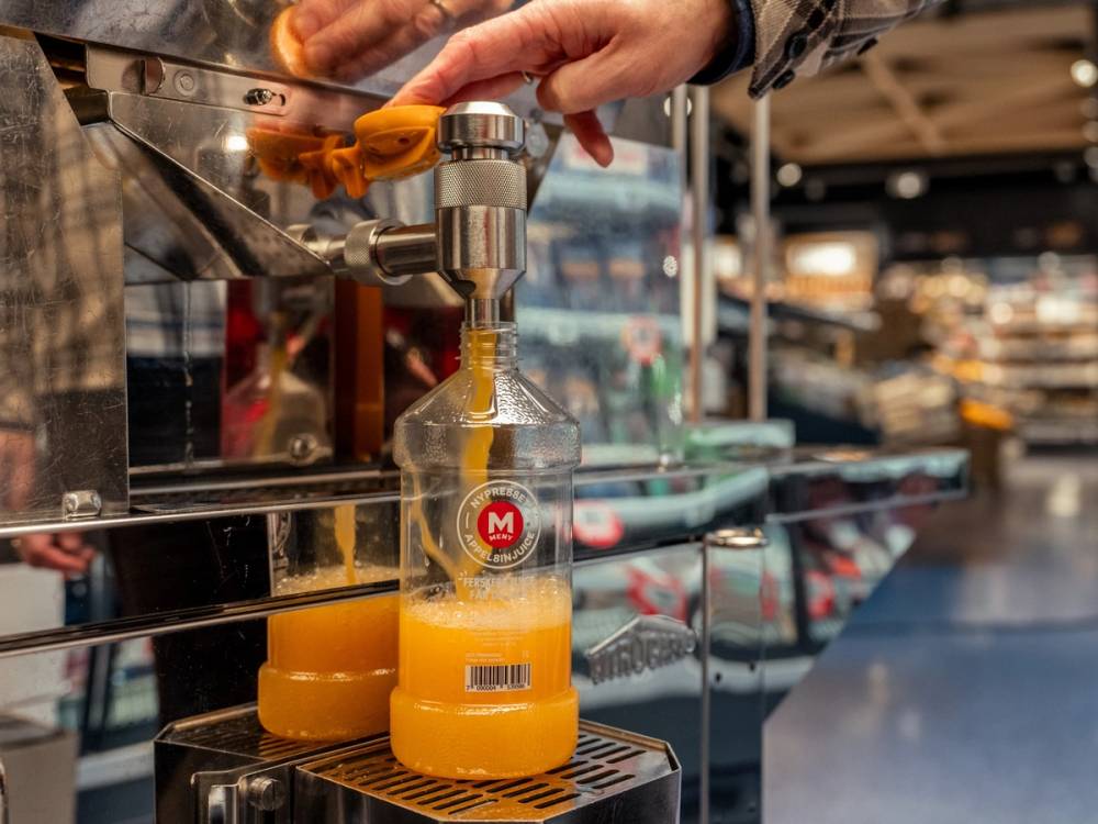 En person heller appelsinjuice fra en flaske i en butikk, med andre flasker og varer i bakgrunnen.