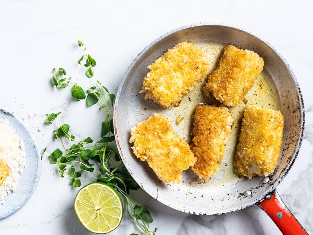 Friterte panerte torskefiskepinner på en tallerken med sitron og urter ved siden av.
