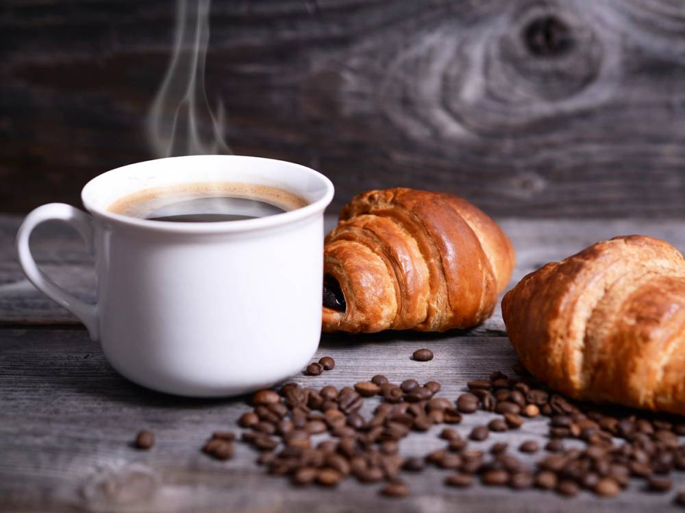 En kopp svart kaffe med to croissanter og kaffebønner på en treplate, mot en mørk trebakgrunn.