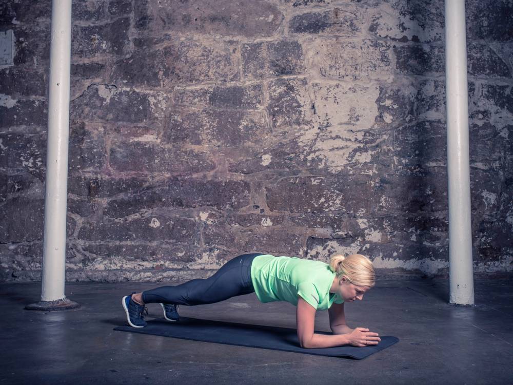 En kvinne gjør en plankeøvelse på gulvet i et treningsrom med en murvegg i bakgrunnen.