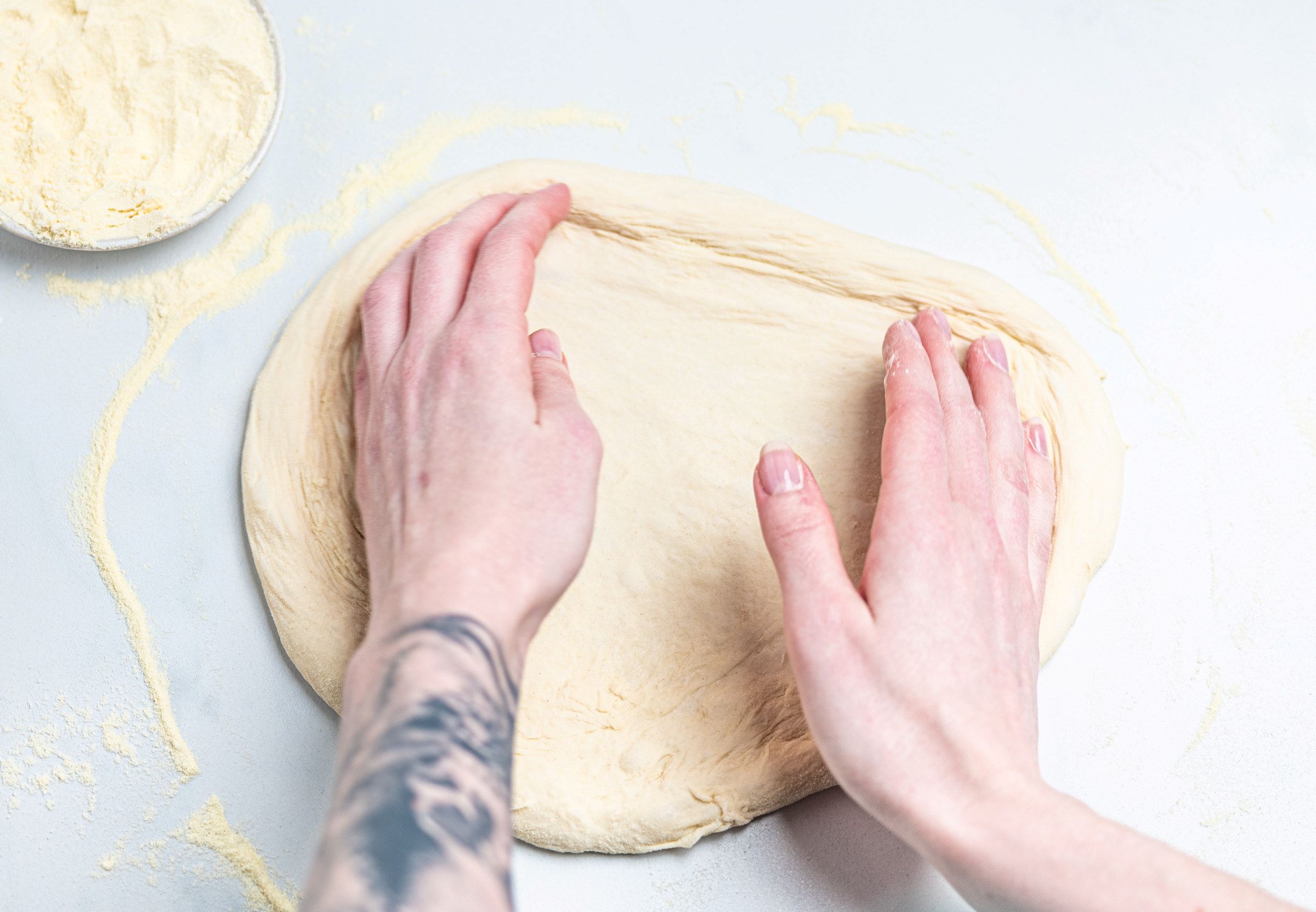 Person som ruller ut pizzadeig med hendene på et hvitt underlag, med en bolle med mel i nærheten.