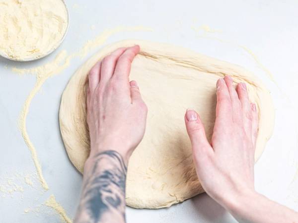 Person som ruller ut pizzadeig med hendene på et hvitt underlag, med en bolle med mel i nærheten.