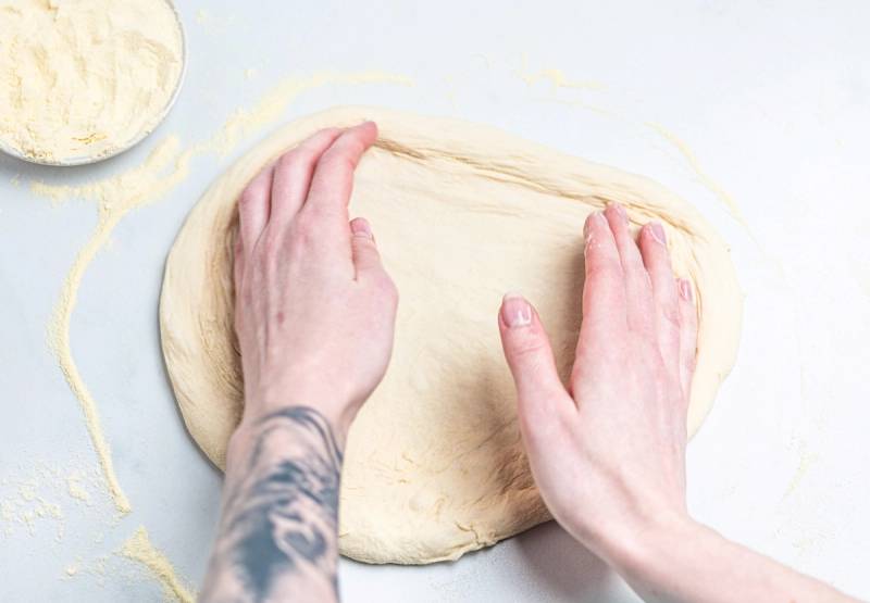 Person som ruller ut pizzadeig med hendene på et hvitt underlag, med en bolle med mel i nærheten.