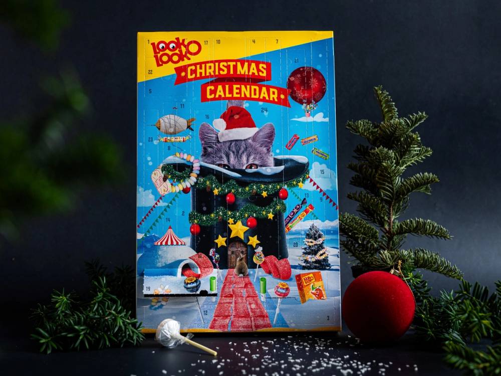 Julekalender for 2023 med en kat som ligger i en peis omgitt av julepynt og pyntede trær, med en rød ball og snø rundt.