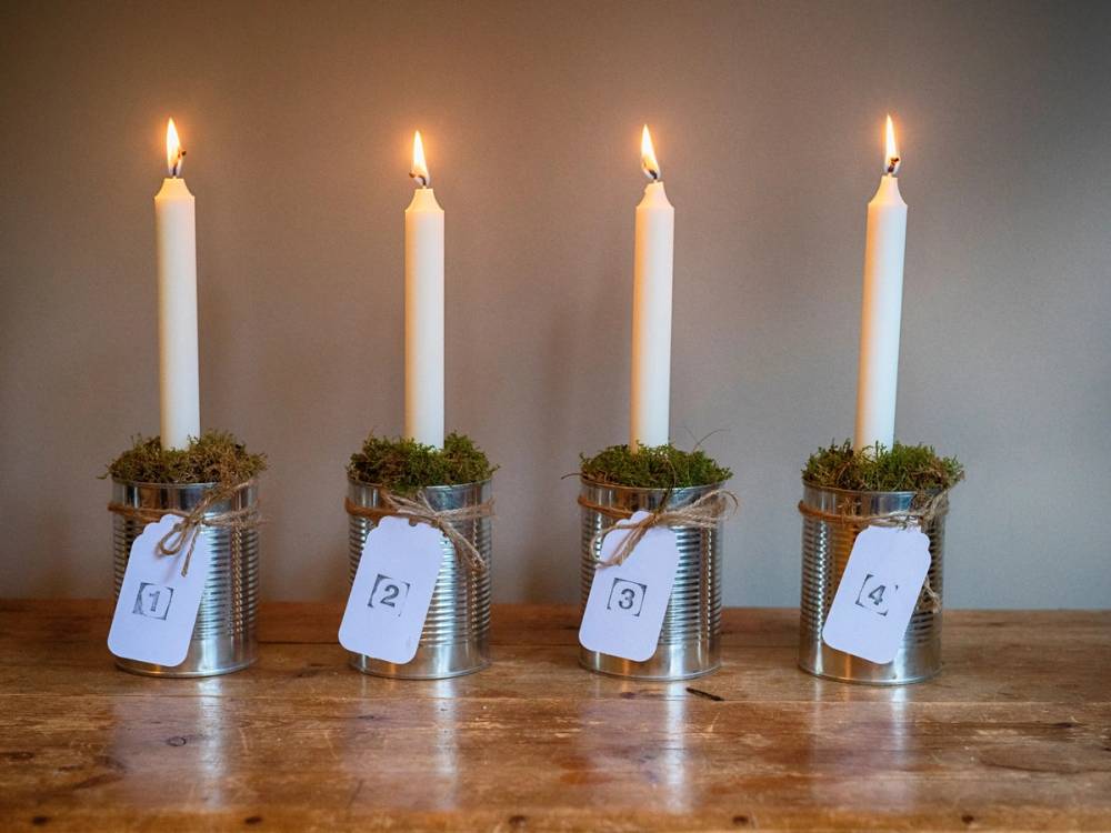 Fire adventsstaker med lysende lys på en trebenet bord, pyntet med små grønne kranser og merkelapper nummerert 1 til 4.