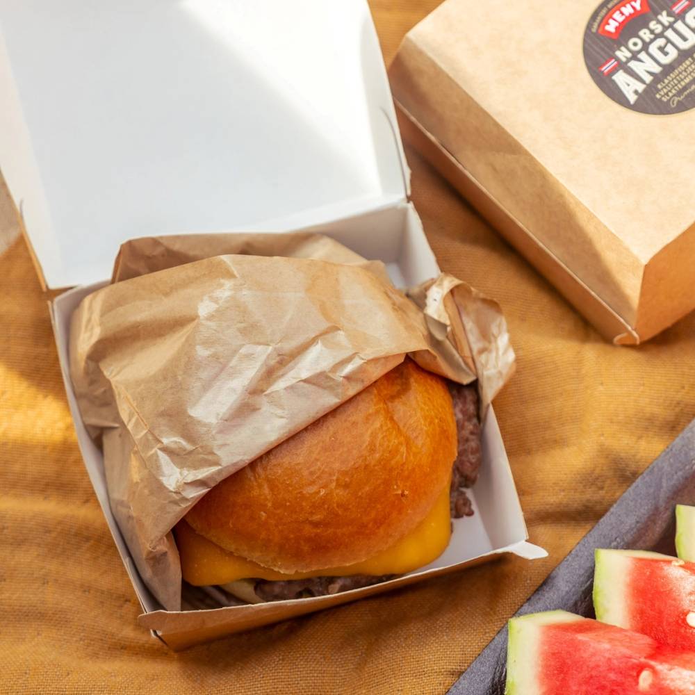En burger i en liten hvit pappeske med brunt papir rundt, sammen med en pappeske med tekst og noen vannmelonskiver på siden.