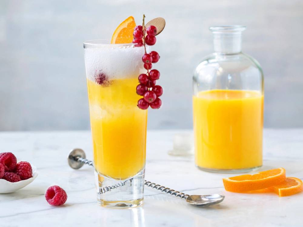 Et glass med appelsinFizz mocktail pyntet med en skive appelsin og røde bær, med en mugge med appelsinjuice i bakgrunnen.