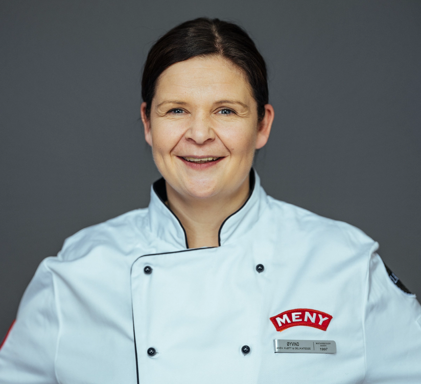 En person i kokkeforkle med MENY-merke, uten synlig ansikt, mot en grå bakgrunn.