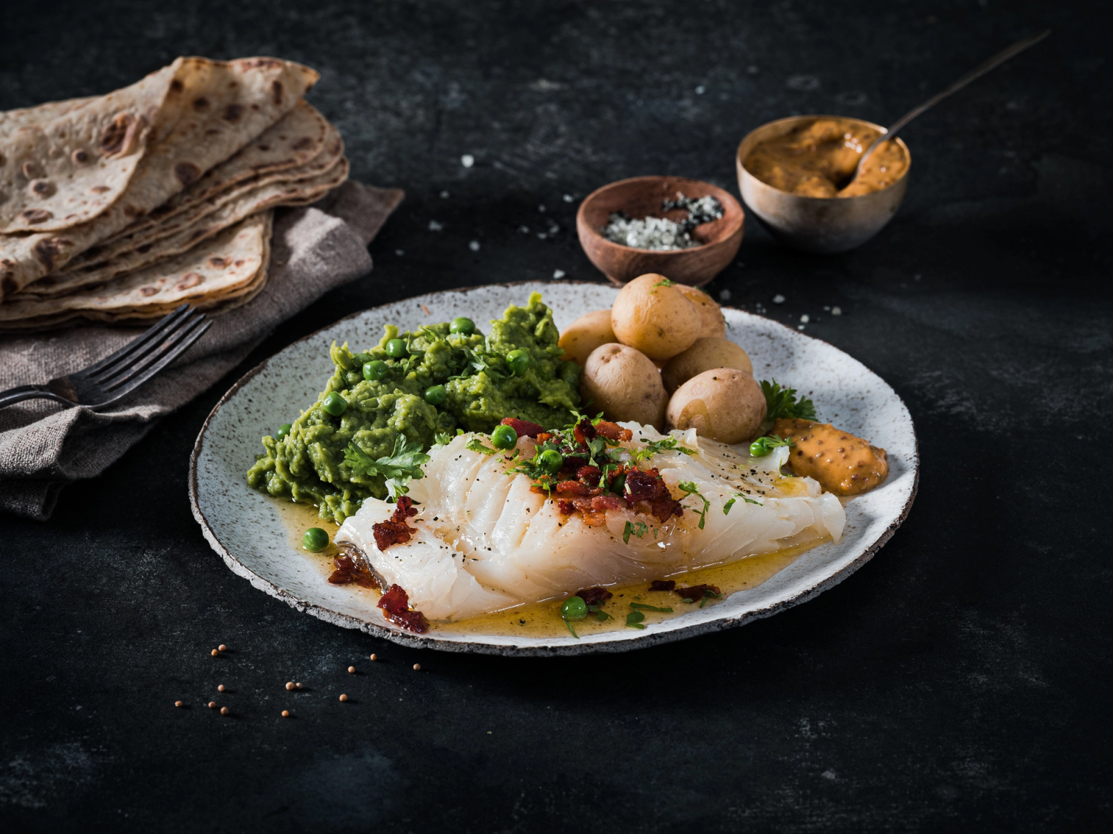 Lutefisk servert med grønnsaker, poteter og tilbehør på en tallerken, med brød og krydder i bakgrunnen.