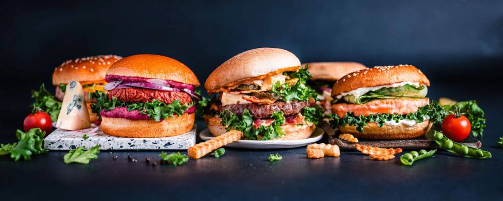 Tre burgere med ulike fyll, omgitt av små tomater, salat og kjeks på et bord.