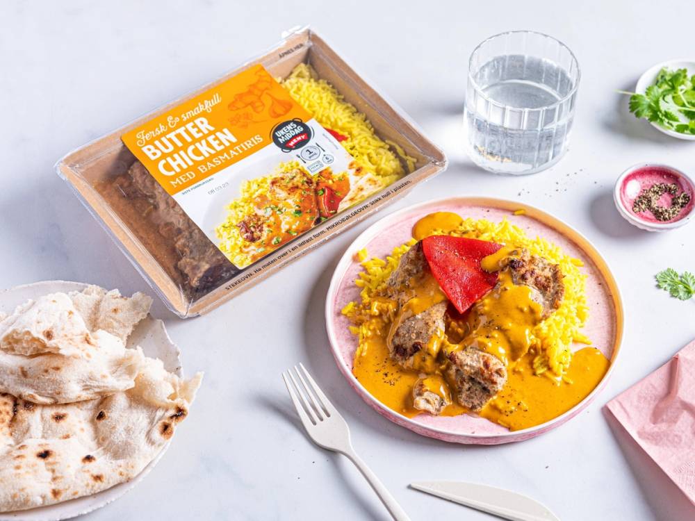 En ferdig oppvarmet takeaway-rett med kylling og ris, servert på en tallerken med en skje ved siden av, omgitt av glass vann og små krydderboller.