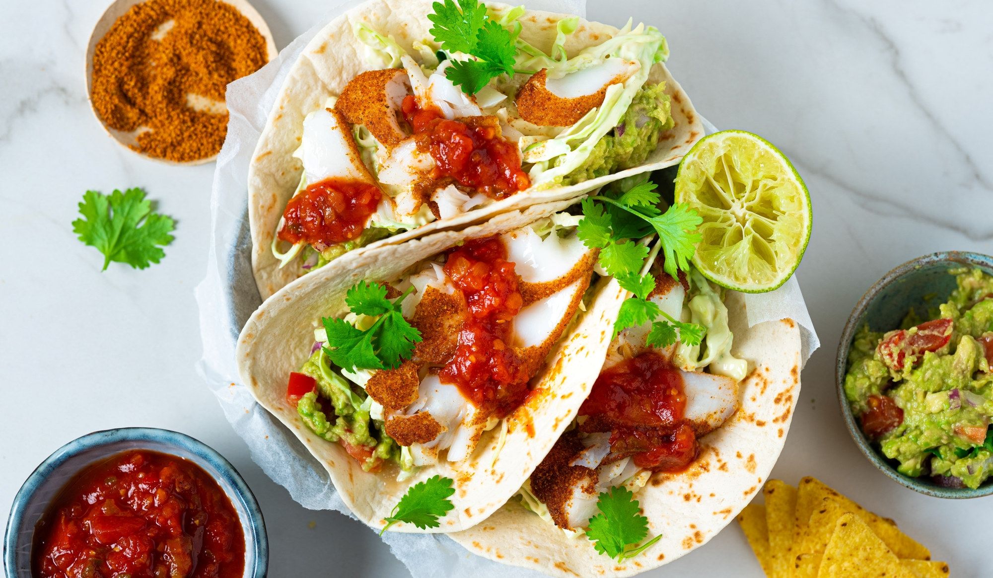 To torsketacos med salat, salsa og sitron på en tallerken, omgitt av små skåler med salsa og guacamole.