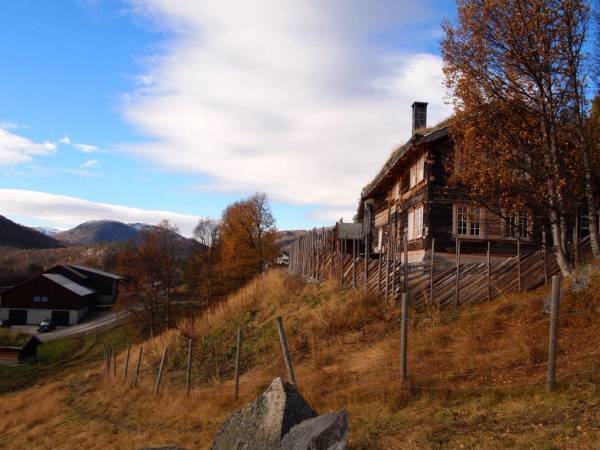 En fjellgård med tradisjonell trebygning omgitt av høstfargede trær og et gjerde, med fjell i bakgrunnen.