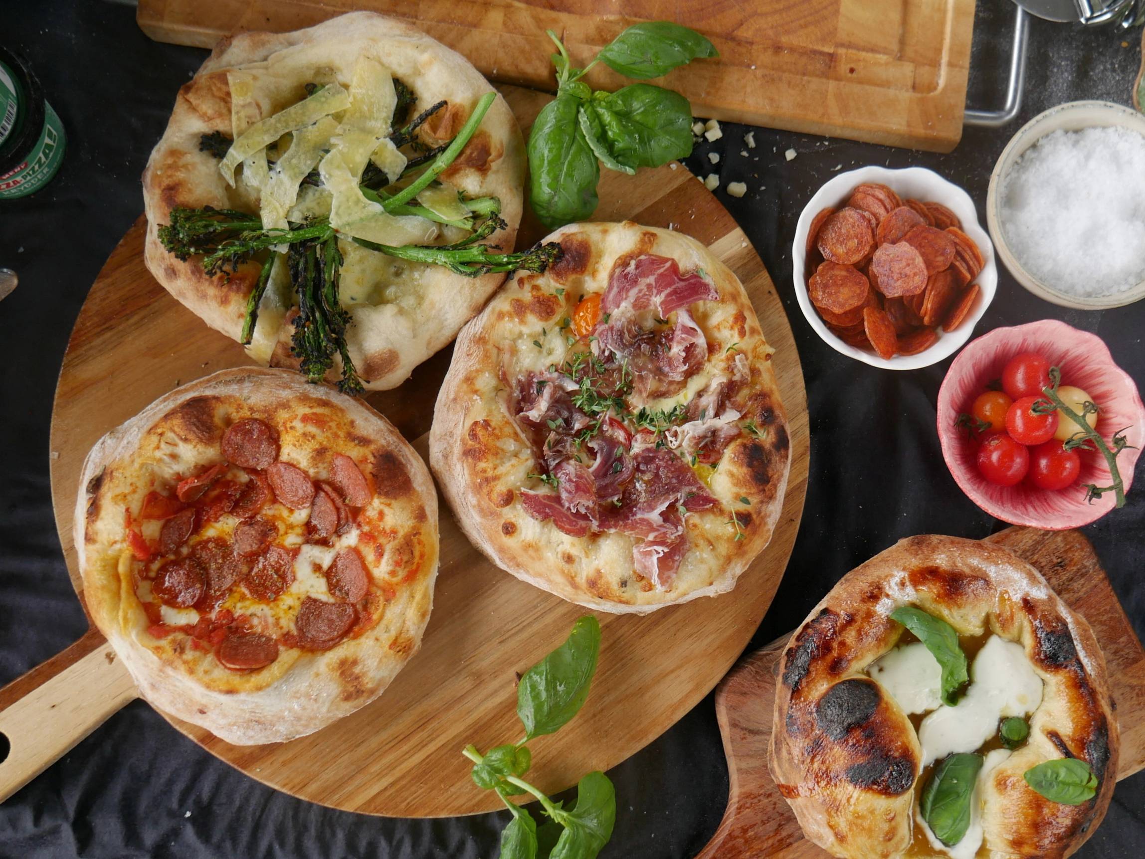 Tre pizzaer med ulike toppings på et rundt trebrett, omgitt av små skåler med tomater, kjøtt og urter.