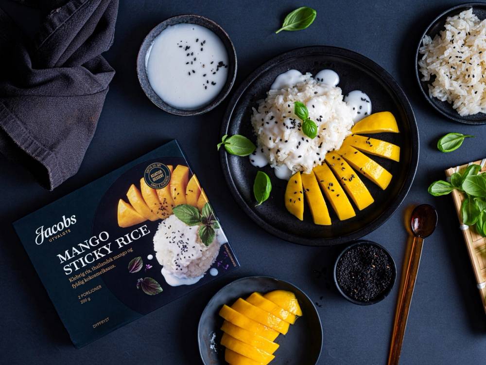 Mango sticky rice servert på en svart tallerken med skiver av mango og en klatt med klebrig ris, omgitt av en kopp kokosmelk og en pakke med mango sticky rice.