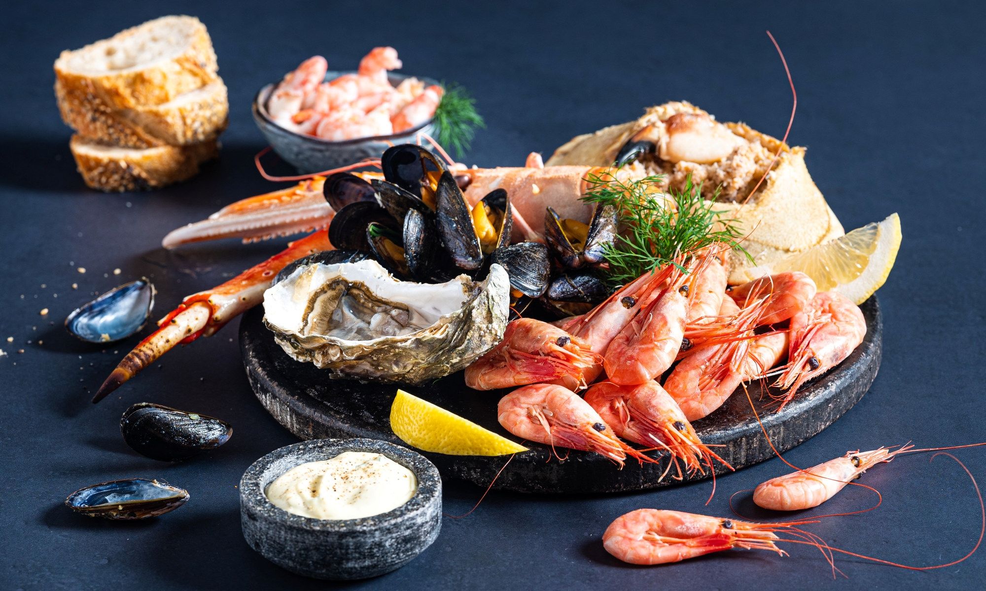 Et utvalg av skalldyr som reker, blåskjell og østers på en steinplate med sitronbåter og dipp, klar for helgekos.