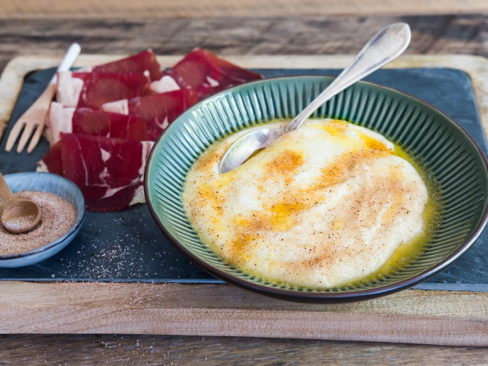 Rommepudding med sukker og en skje i en grønn skål, med et stykke rosinbrød på en tallerken i bakgrunnen