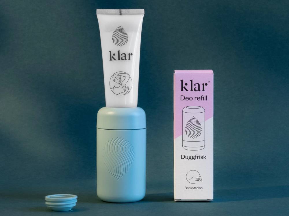 En deodorantflaske med tilhørende eske står på en blå bakgrunn, med en liten blå beholder ved siden av.