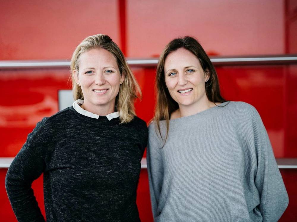 To kvinner står side om side foran en rød bakgrunn, smilende og kledd i casual klær.