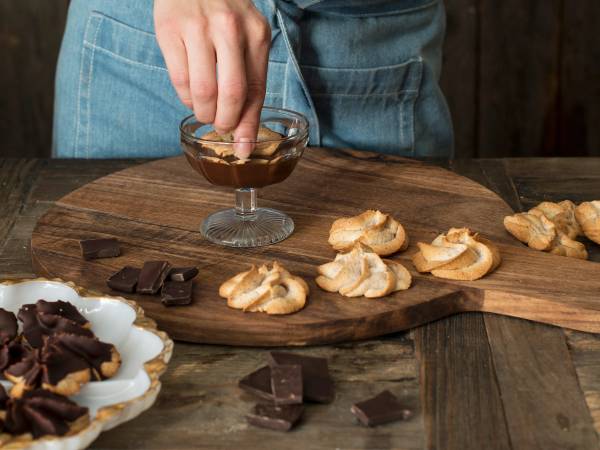 Person som pynter kransekaketopper med sjokolade og krem på et trefat, med flere kransekaketopper og sjokoladebiter rundt.