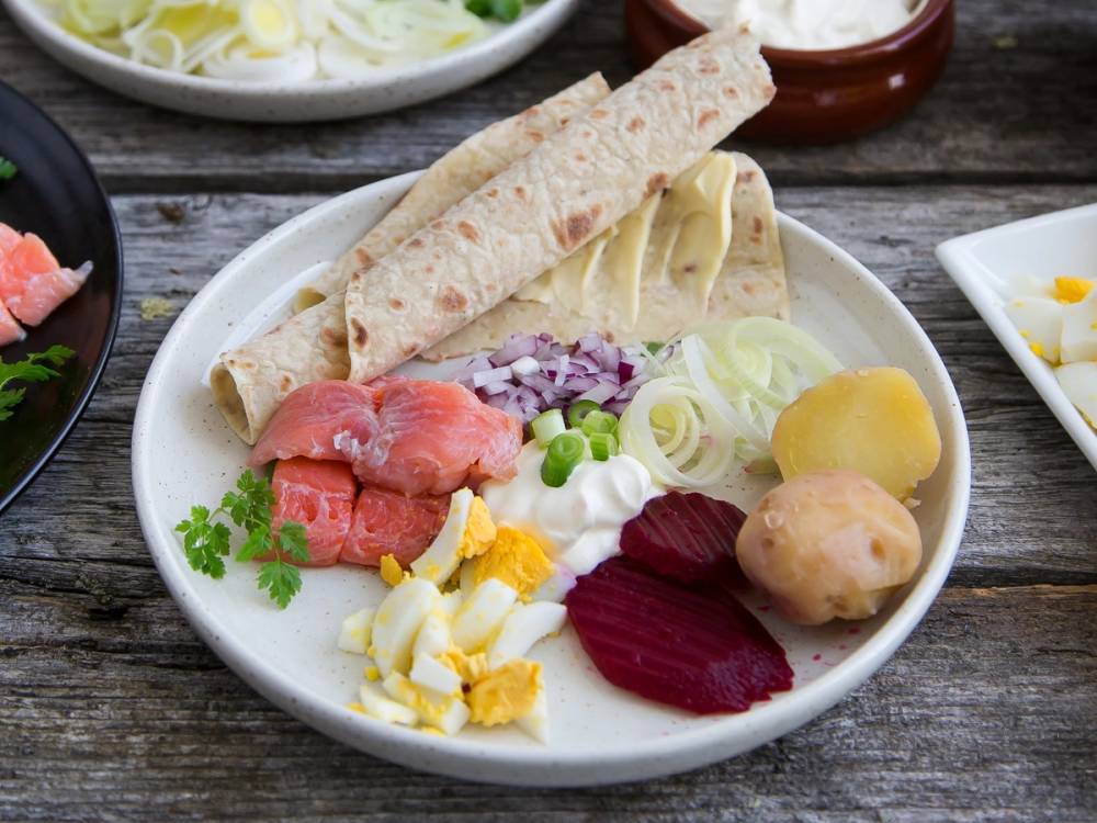 Tradisjonell rakfisk servert med flatbrød, løk, rømme, poteter og grønnsaker på en hvit tallerken.