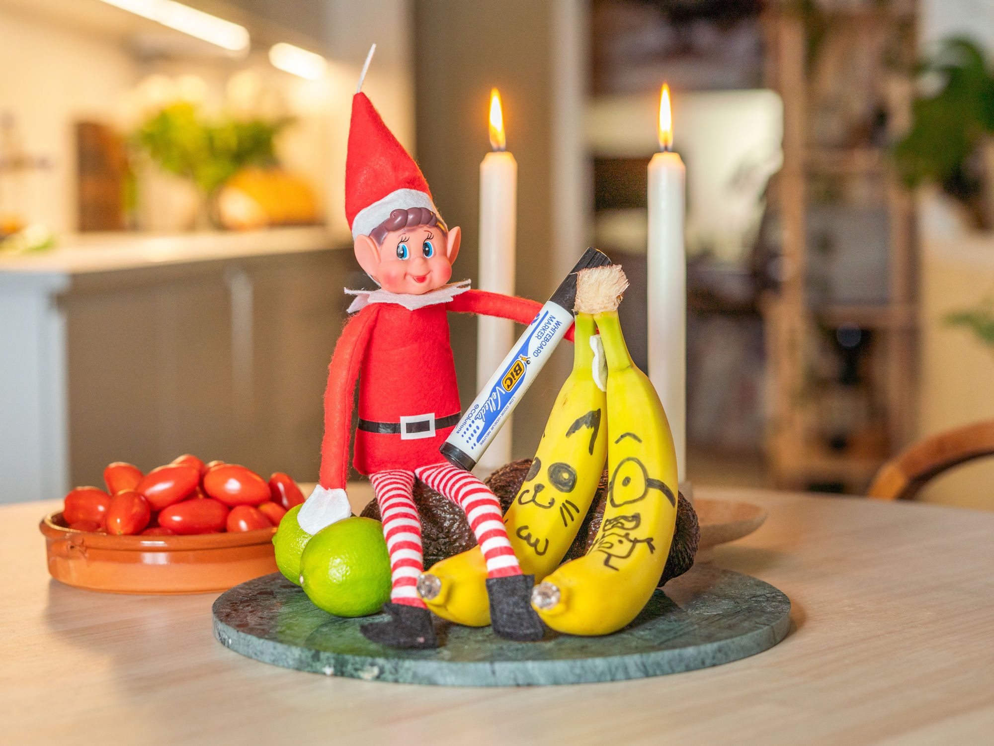 En julepyntet rampenisse med rød lue og stripete klær sitter på et bord sammen med bananer, en grønn eple og et fat med tomater, omgitt av julelys og stearinlys.