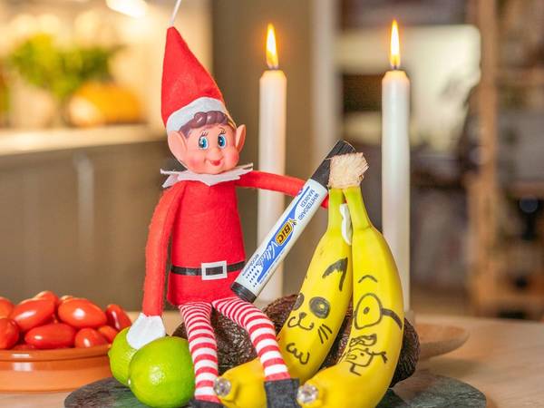 En julepyntet rampenisse med rød lue og stripete klær sitter på et bord sammen med bananer, en grønn eple og et fat med tomater, omgitt av julelys og stearinlys.