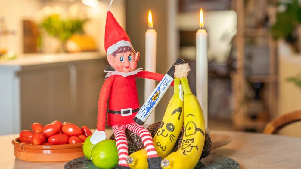 En julepyntet rampenisse med rød lue og stripete klær sitter på et bord sammen med bananer, en grønn eple og et fat med tomater, omgitt av julelys og stearinlys.