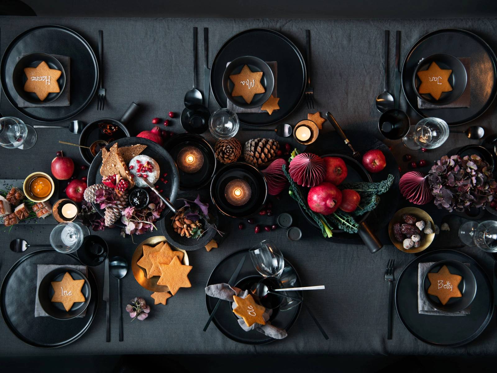 Dekket julebord med tallerkener, lys, julepynt og småkaker på et mørkt bord.
