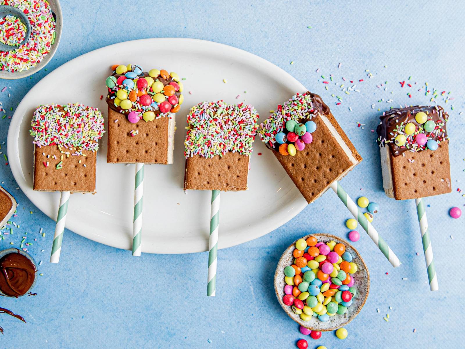 Fire iskrem-sandwicher med sjokoladekjeks og fargerike nonstop på en hvit tallerken, omgitt av små godisbiter og en skje med godteri.