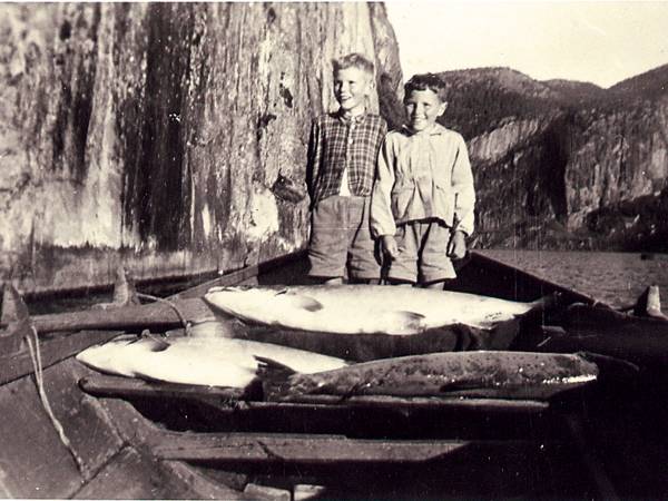 To barn står i en liten båt med fisk, omgitt av fjell og vann, på et gammelt fiskebilde.