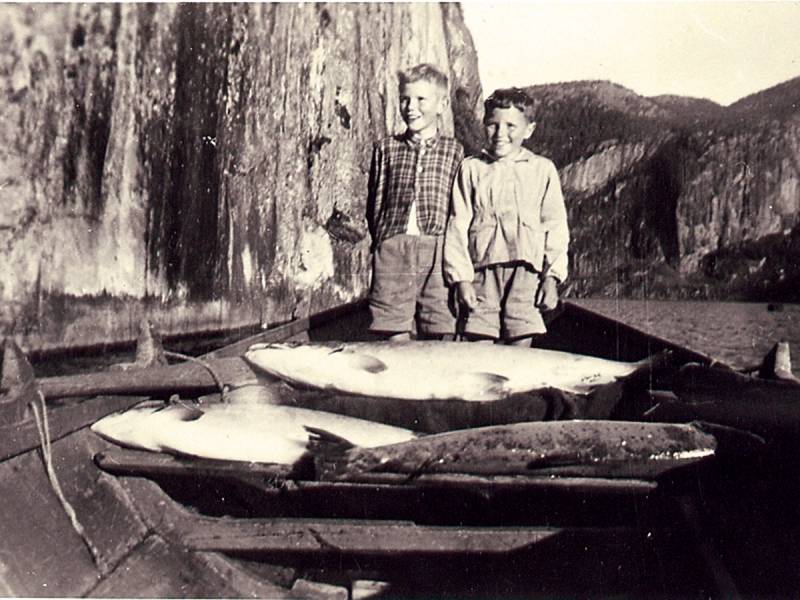 To barn står i en liten båt med fisk, omgitt av fjell og vann, på et gammelt fiskebilde.