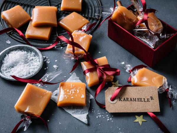 Fløtekarameller pakket inn i cellofan og pyntet med røde bånd, lagt på en mørk overflate med litt melis og en liten skål med melis ved siden av.