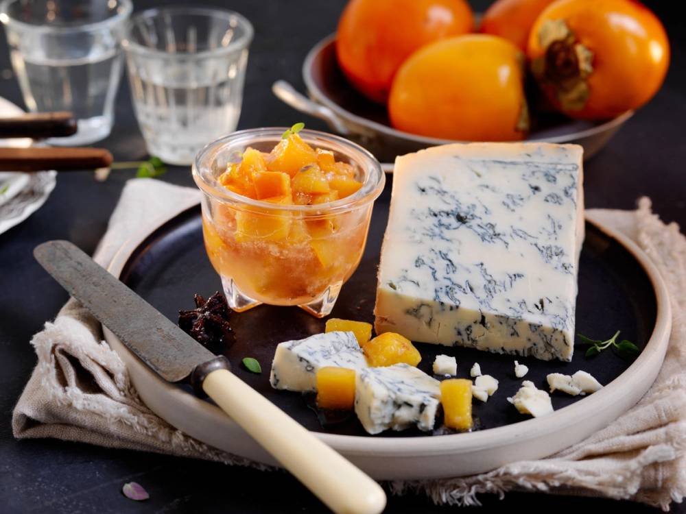Persimon syltetøy servert sammen med blåmuggost og noen skiver av ost, med et fat med persimonfrukter i bakgrunnen.