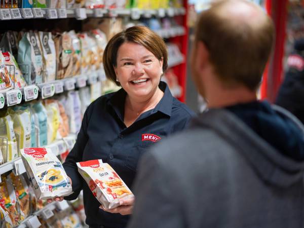 En kvinne i butikk holder en pakke med melk og snakker med en mann med ryggen til, i en dagligvarebutikk.