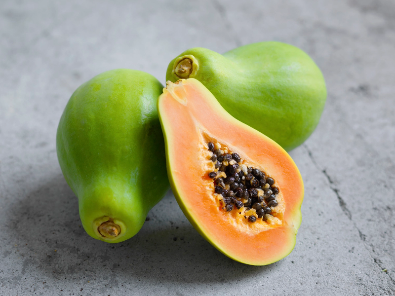To hele papayaer og en skåret papaya med oransje fruktkjøtt og svarte frø, plassert på en grå overflate.