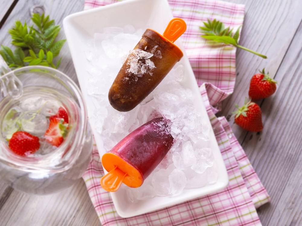 To hjemmelagde iskrembarer med trepinner på en hvit serveringsplate, omgitt av jordbær og isbiter, på et rosa og hvitt rutete kjøkkenhåndkle.