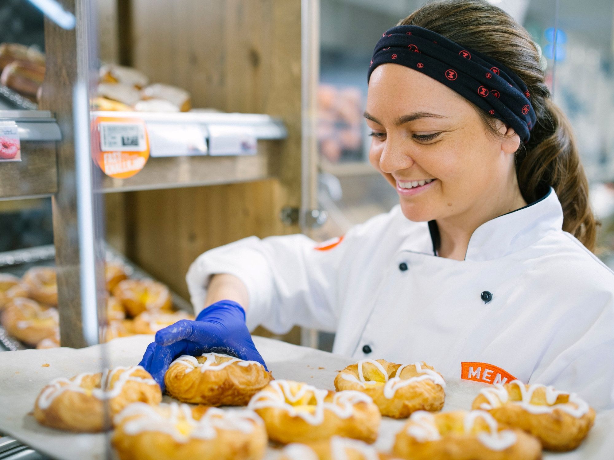 En person i hvit kokkeuniform og blå hansker jobber med å pakke eller sortere bakverk i en butikk eller på et bakeri.