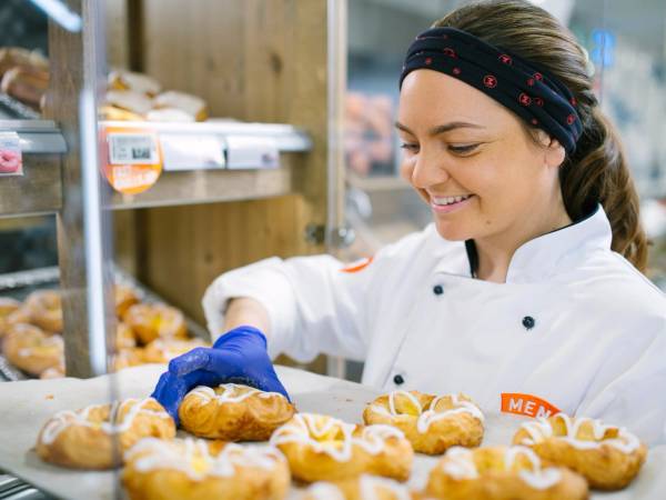 En person i hvit kokkeuniform og blå hansker jobber med å pakke eller sortere bakverk i en butikk eller på et bakeri.