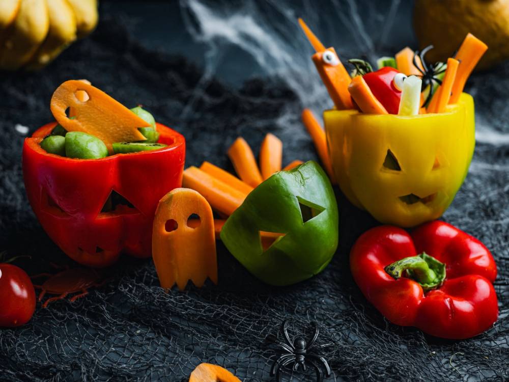 To gule og en rød pepper med skåler av frukt og grønnsaker, dekorert til Halloween, med små ansikter og spiselige elementer rundt på en svart bakgrunn.