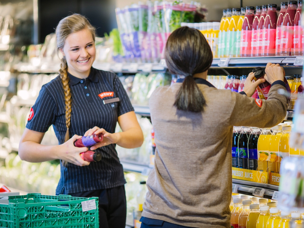 To ansatte fyller ut varer sammen i en dagligvarebutikk, en av dem har langt hår i fletter og en annen har kort hår og bruker en grå genser.