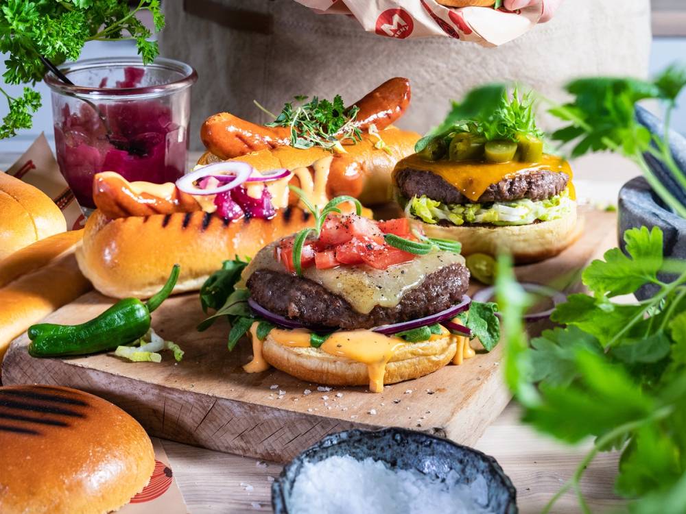 Et utvalg av grillmat og tilbehør, inkludert burgere, pølser, grønnsaker og saus, på et bord.