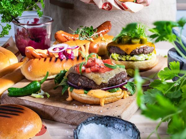 Et utvalg av grillmat og tilbehør, inkludert burgere, pølser, grønnsaker og saus, på et bord.