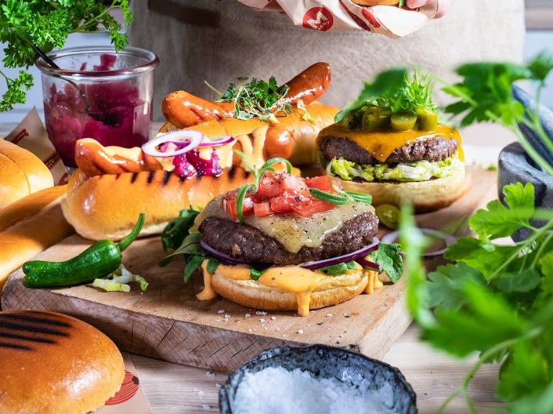 Et utvalg av grillmat og tilbehør, inkludert burgere, pølser, grønnsaker og saus, på et bord.