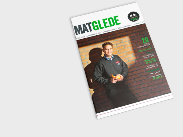 Forside av MatGlede med to personer som smiler og holder mat, mot en murvegg, med grønn og hvit tekst.