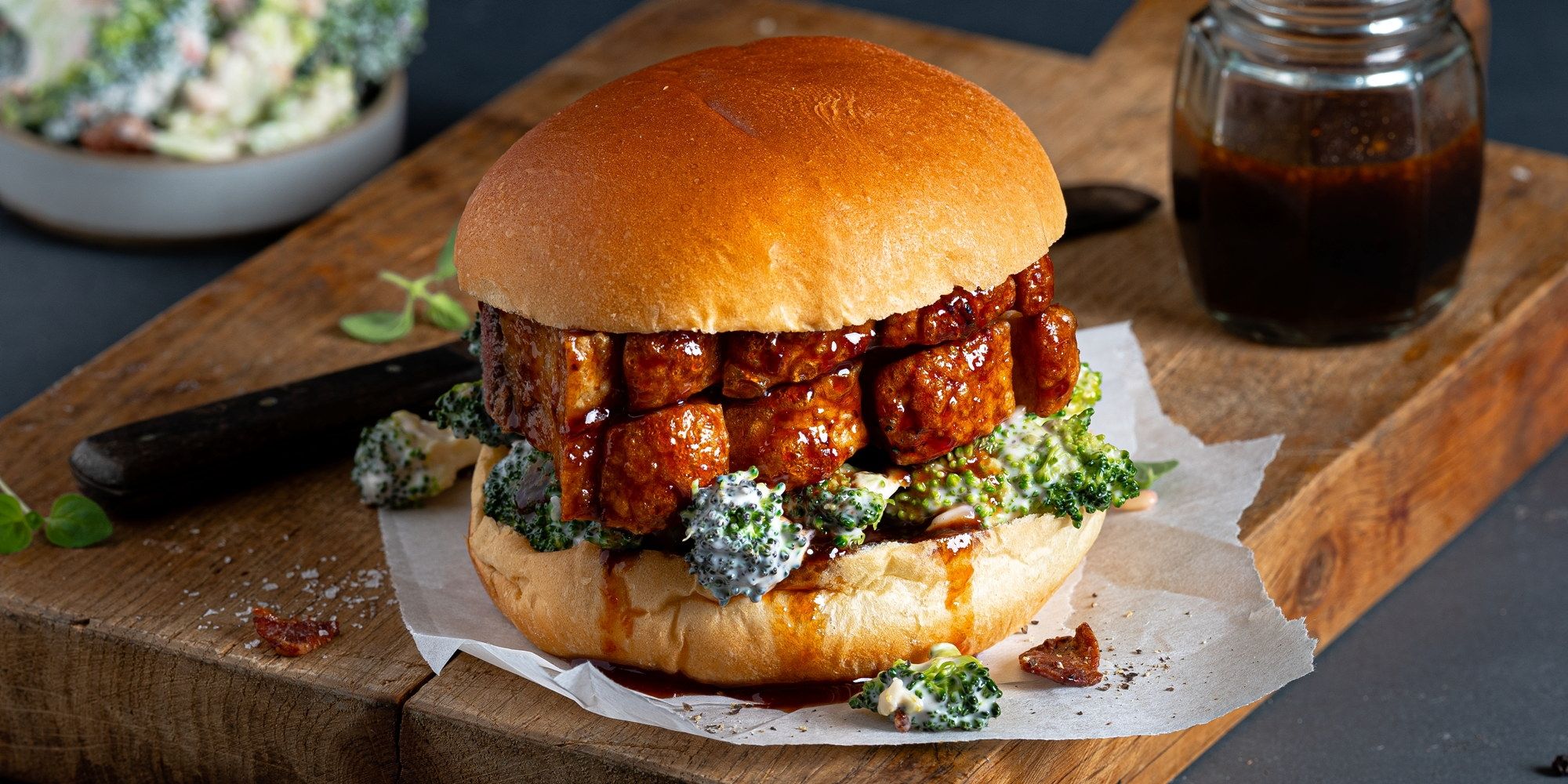 Ribbesandwich med broccolisalat på en treplate, med en liten glass med saus i bakgrunnen.