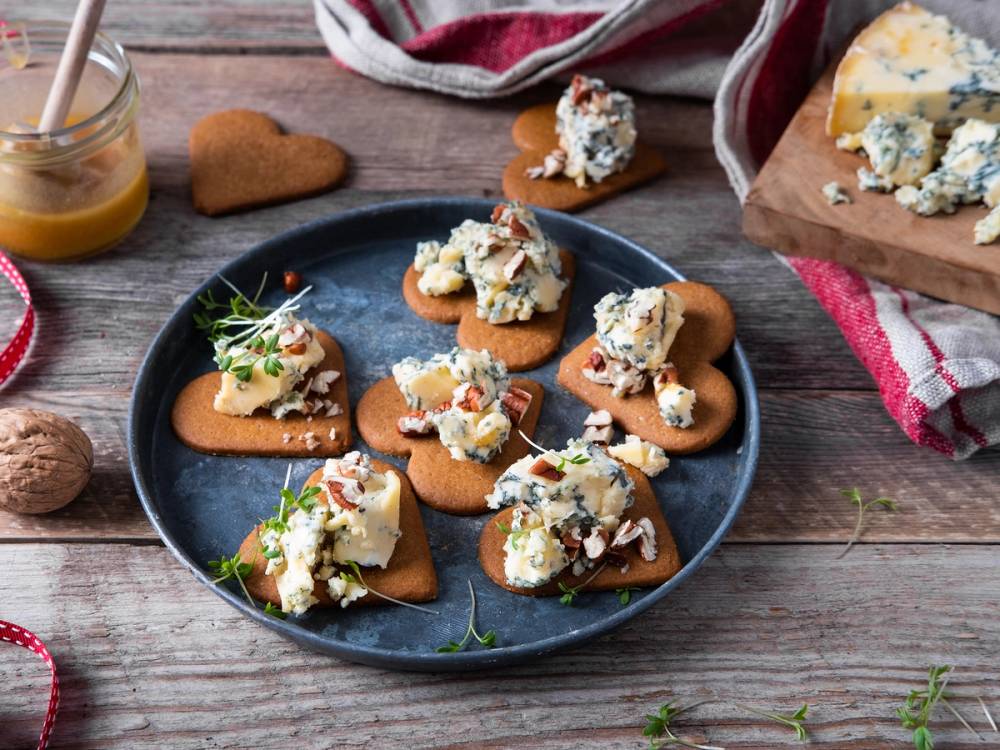 Pepperkaker med blåmuggost og urter på en blå tallerken, omgitt av andre småkaker og tilbehør på et trebord.